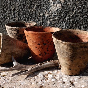 Könnte beinhalten: Eine Sammlung verwitterter Terrakotta-Töpfe in verschiedenen Größen und Brauntönen. Die Töpfe sind auf einer Steinoberfläche mit einem verwitterten Geweih angeordnet. Der Hintergrund ist eine dunkle, strukturierte Wand.
