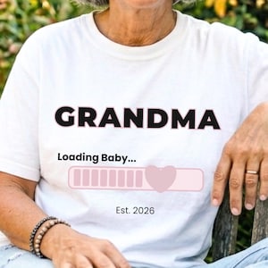 Puede incluir: Una camiseta blanca de manga corta con el texto negro GRANDMA, seguido del texto Loading Baby... sobre una barra de progreso rosa con un icono de corazón, y el texto Est. 2026 en la parte inferior. La camiseta se lleva con vaqueros azul claro.