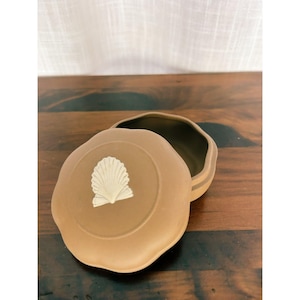 May include: A round, matte tan ceramic trinket box with a removable lid. The lid features a raised, white, fan-shaped seashell relief centered on its surface. The container has a smooth, uniform finish and a simple, circular silhouette.