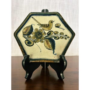 May include: A hexagonal ceramic tile featuring a hand-painted design of a bird perched on a large flower with leafy stems. The tile has a cream-coloured background with dark blue and brown painted details and a dark blue border on the edges.