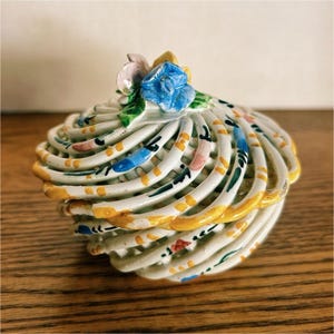 May include: A decorative ceramic trinket box with a spiral design. The box is white with yellow, blue, pink, and green accents. A blue rose and other flowers adorn the lid. The box sits on a wooden surface.