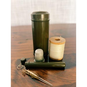 May include: A sewing kit containing a tall olive green cylindrical container with a lid, a smaller matching green tube, a silver metal thimble, a wooden spool of white thread, and two sewing needles with thread wrapped around their ends.