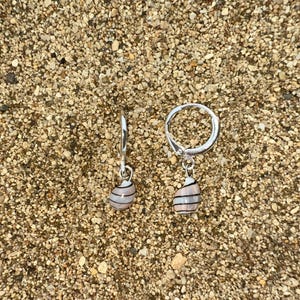 May include: A pair of silver hoop earrings with seashell charms. Each charm is a light pink color with black stripes. The earrings are displayed on a sandy surface.
