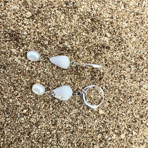 May include: A pair of earrings with a pearl and a white shell-shaped pendant. The earrings are silver-toned and rest on a bed of sand. One earring has a hoop closure, while the other has a hook.