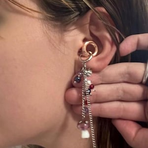 May include: A rose gold-toned ear cuff featuring a dangling silver-toned chain attachment. The chain is adorned with small, multi-colored beads and a miniature mushroom charm with a purple cap and white stem, creating a layered, dangling aesthetic.