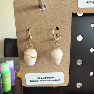 Puede incluir: Un par de pendientes con conchas marinas hawaianas naturales suspendidas de aros chapados en oro de 18 quilates. Las conchas son de color crema y bronceado con una forma lisa y cónica. La etiqueta dice 18k gold plated Natural hawaiian seashell.