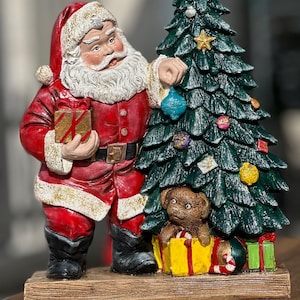 May include: A Christmas figurine of Santa Claus in a red suit, holding a gift and decorating a green Christmas tree. A small teddy bear is in a yellow gift box. Ornaments and gifts are at the base. The figurine is on a wooden base.
