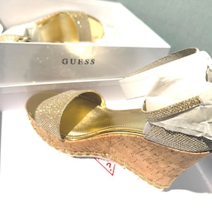 Puede incluir: Sandalia dorada con cuña, parte superior brillante y suela de corcho, presentada en una caja blanca con la marca "GUESS". La sandalia tiene una correa en el tobillo y está parcialmente envuelta en papel de seda blanco.