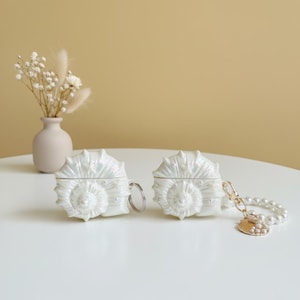 May include: Two white, iridescent seashell-shaped AirPod cases with silver key rings. One case has a pearl bracelet and a gold charm. A small vase with dried flowers sits in the background. The cases are on a white surface.