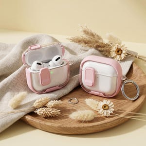 Puede incluir: Dos fundas protectoras rosas y transparentes para auriculares inalámbricos. Una funda está abierta, revelando auriculares blancos. Las fundas tienen un marco rosa y un llavero plateado. Las fundas están sobre una bandeja de madera con flores secas.