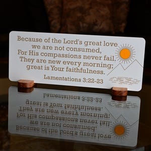 May include: A lit white candle on a decorative plate, next to a sign with the text "Because of the Lord's great love..." supported by wooden blocks. The sign features a sun and mountain graphic. The candle and sign are reflected.