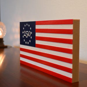 Könnte beinhalten: Eine amerikanische Flagge aus Holz mit roten und weißen Streifen und einem blauen Kanton mit weißen Sternen und der Zahl "250". Die Flagge wird auf einem Holzblock präsentiert, der ein patriotisches Thema verkörpert.