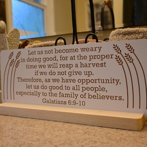 May include: A white sign with brown text, quoting Galatians 6:9-10. The sign is supported by a light wood base and has wheat stalk designs. An angel figurine and a woven basket are in the background.
