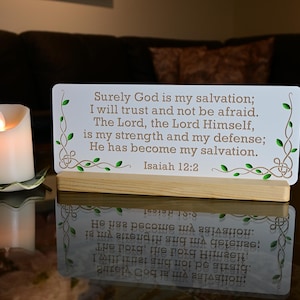 May include: A white sign with the text "Surely God is my salvation" and a Bible verse, displayed on a wooden stand. A lit, white candle sits to the left on a decorative holder. The scene is reflected on a glass surface.