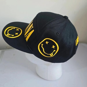 May include: A black baseball cap featuring bright yellow graphics. The design includes a yellow smiley face with X-shaped eyes and a tongue sticking out on the side and brim, along with the yellow text NIRVANA printed on the front panel.