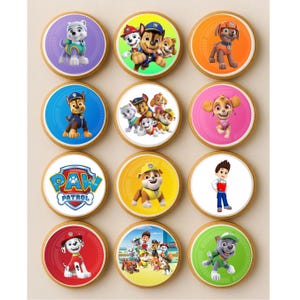 May include: A dozen round cookies with colorful images of characters from the animated series Paw Patrol. Each cookie has a white border and a golden-brown base. The images include the Paw Patrol logo and various characters like Chase, Marshall, and Skye.
