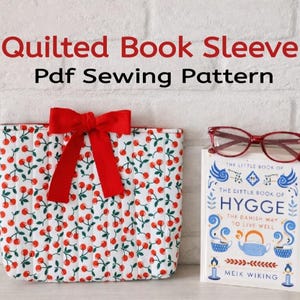 May include: A quilted book sleeve with a cherry pattern and a red bow. The book sleeve is next to a book titled "The Little Book of Hygge" and a pair of red glasses. The text "Quilted Book Sleeve Pdf Sewing Pattern" is at the top.