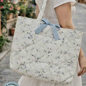 以下が含まれることがあります： 白と青の花柄のキルティングトートバッグ。バッグには水色のリボンと前面にポケットが付いています。バッグは、同じ素材で作られたストラップで肩からかけています。