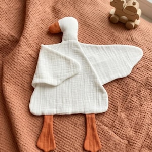 May include: A white goose-shaped baby comforter with orange feet and beak, lying on a textured peach-colored blanket. A small wooden camel toy is in the background. The comforter is made of soft, textured fabric.