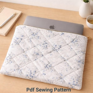 Pode incluir: Uma capa acolchoada branca para laptop com um padrão floral azul. A capa está aberta, revelando um laptop cinza com o logotipo da Apple. O zíper é dourado e o texto "Pdf Sewing Pattern" está na parte inferior.