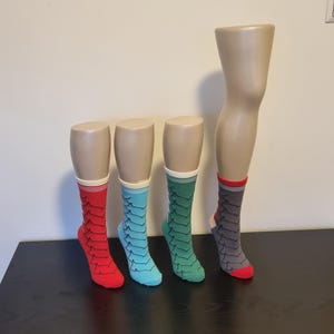 Op de afbeelding: Vier paar sokken met patronen die op mannequinbenen worden getoond. De sokken hebben een schubachtig ontwerp in rood, blauwgroen, groen en grijs, elk met een contrasterende manchet en teen. De sokken worden tentoongesteld op een zwart oppervlak tegen een neutrale muur.