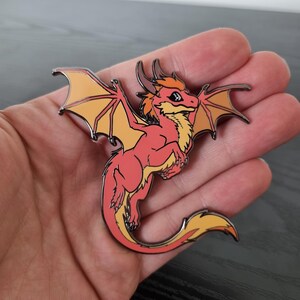 Op de afbeelding: Een harde emaille pin met een kleine, gestileerde draak met koraalroze schubben, een crèmekleurige buik en oranje getinte vleugels. De draak heeft twee kleine hoorns, een lange gebogen staart en is omlijnd met een donkere metalen afwerking.