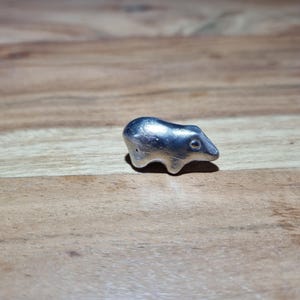 Op de afbeelding: Een klein, zilverkleurig metalen beeldje in de vorm van een gestileerde beer. Het object heeft een glad, reflecterend oppervlak met een afgerond lichaam, vier korte poten en een kop met een zichtbaar rond oogdetail.