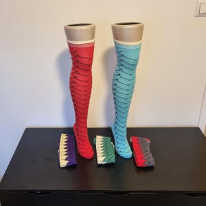 May include: Two pairs of thigh-high stockings, one red and one turquoise, displayed on mannequins. Each stocking features a scale-like pattern. Three pairs of folded socks with similar patterns in purple, green, and red are also visible.