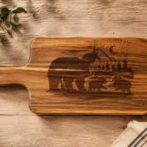 May include: A rectangular wooden cutting board with a handle featuring a laser-engraved design of a large bear and two cubs in a forest setting with mountains and a crescent moon in the background. The board has a natural brown wood grain finish.