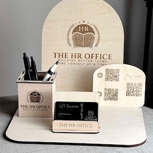 May include: A wooden desk organizer set with the text "THE HR OFFICE" and a logo. The set includes a pen holder, a business card holder, and a display stand with QR codes. The pen holder contains pens and a marker.