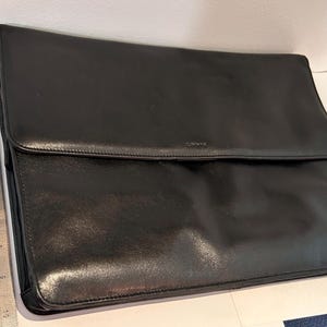 May include: A black leather portfolio case with a fold-over flap closure. A metal ruler is positioned vertically along the left side, showing the case measures approximately 11 inches in height and 15 inches in width across the bottom edge.