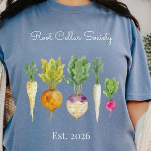 May include: A blue t-shirt featuring a watercolor illustration of root vegetables, including parsnips, beets, and turnips. The text "Root Cellar Society" is arched above the vegetables, with "Est. 2026" below. The shirt is a comfortable, casual style.