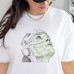 May include: White t-shirt with a detailed graphic of a futuristic face with a helmet and visor. The design is in shades of green and white, with fine lines and shading. The person is wearing a black and white trucker hat.