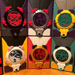 Peut inclure: Une collection de dix boîtes géométriques colorées imprimées en 3D. Chaque boîte présente un design circulaire avec une illustration de personnage unique au centre. Les boîtes sont de couleurs variées, dont le jaune, le rouge, le noir et le violet.