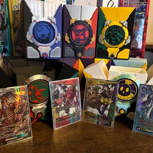 May include: A collection of colorful Digimon card game accessories and cards. The image features several card holders in blue, red, and yellow, along with various Digimon cards. The cards have detailed illustrations and text, and the card holders have circular designs.