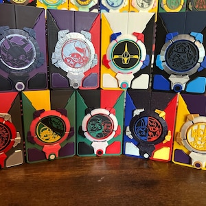 May include: A collection of rectangular plastic cases featuring circular emblems with various character designs. Each case displays a unique colour scheme, including purple, red, yellow, blue, green, and black, with intricate moulded details.