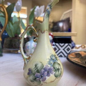 May include: A small, hand-painted porcelain pitcher with a curved handle and spout. The pitcher is primarily cream-colored, adorned with purple and blue floral designs and green leaves. Gold accents trim the handle and base, adding a touch of elegance.