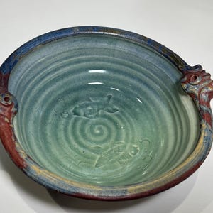 May include: Handmade ceramic bowl with a fish design. The bowl features a gradient of blue and green hues, with fish and bubbles embossed on the interior. The rim is a darker blue, and the handles are red and blue.