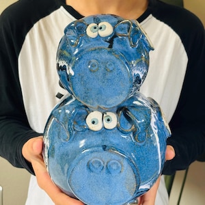 May include: Two stacked ceramic pig sculptures in shades of blue. The pigs have large, expressive eyes and are glazed with a glossy finish. The sculptures are handmade and have a whimsical, artistic design.