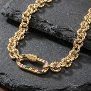 May include: Gold-toned chain necklace with a textured link design. The necklace features a large, decorative clasp with colorful rectangular gemstones in green, orange, and purple. The jewelry is displayed on a dark, textured surface.