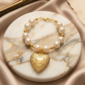 May include: A gold-toned bracelet featuring a series of round white pearls interspersed with small gold beads. A large, heart-shaped gold locket with intricate floral engraving and a smooth central heart detail hangs from the center of the bracelet.