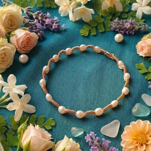 May include: A handcrafted bracelet featuring a series of white pearls strung on a copper wire. The bracelet is displayed on a teal surface, surrounded by flowers, including roses, jasmine, and lavender. The overall aesthetic is delicate and feminine.