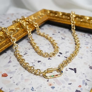May include: A gold-colored chain necklace with a large clasp featuring colorful gemstones. The necklace is made of interlocking oval links. The clasp has a spring-loaded mechanism. The necklace is displayed on a patterned surface.