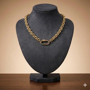May include: A gold-colored chain necklace with a textured design and a decorative clasp. The clasp features green, red, and white accents. The necklace is displayed on a black mannequin bust.