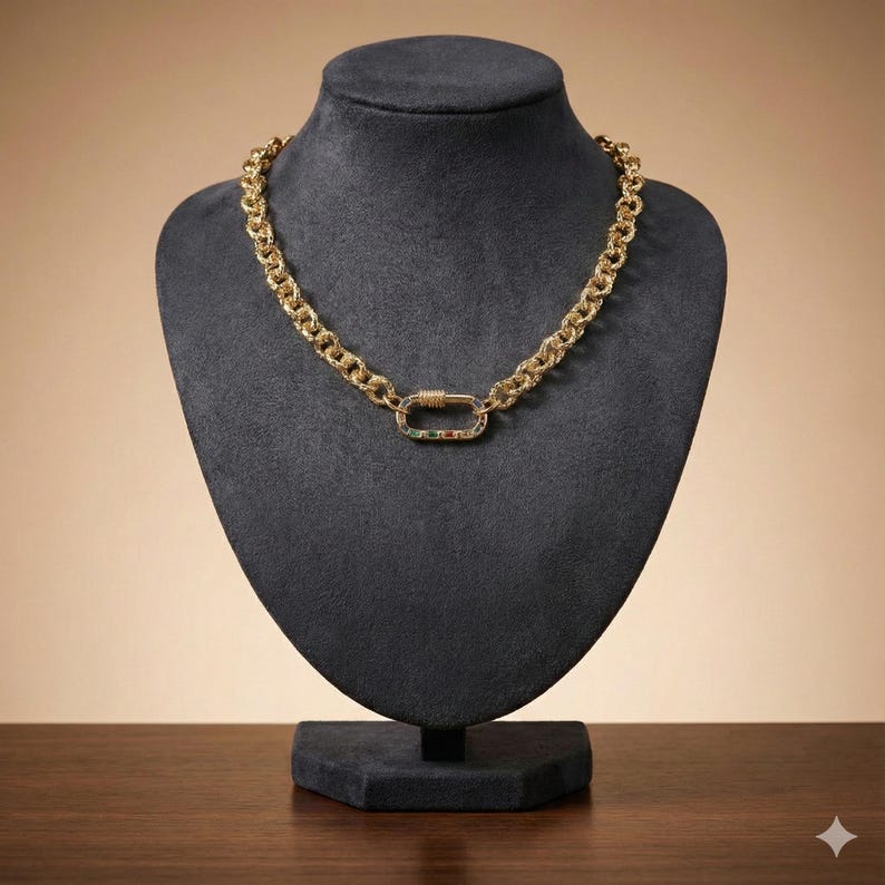 May include: A gold-colored chain necklace with a textured design and a decorative clasp. The clasp features green, red, and white accents. The necklace is displayed on a black mannequin bust.