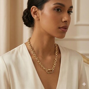 May include: A gold-colored chain necklace with a large oval clasp. The necklace is made of round links and is worn around the neck. The clasp has a colorful design.