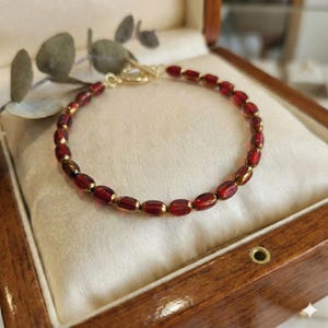 May include: A gold-toned bracelet with oval, deep red beads. The beads are strung together with gold-colored links. The bracelet is displayed inside a wooden jewelry box with a cream-colored interior.
