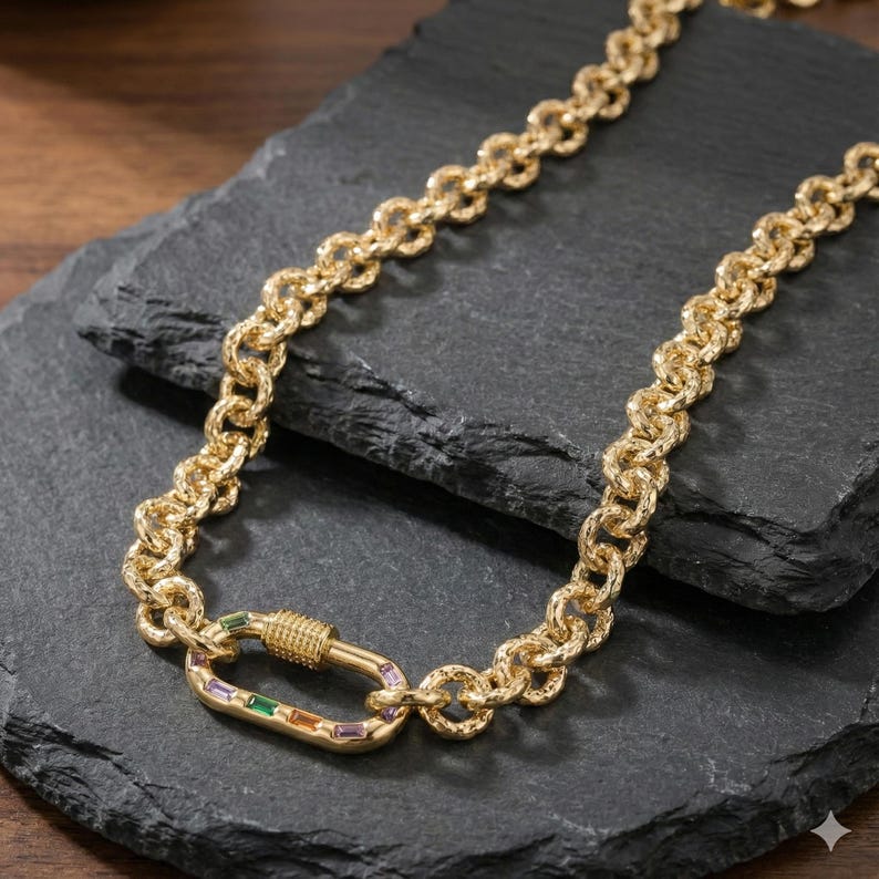 May include: Gold-toned chain necklace with a textured link design. The necklace features a large, decorative clasp with colorful rectangular gemstones in green, orange, and purple. The jewelry is displayed on a dark, textured surface.