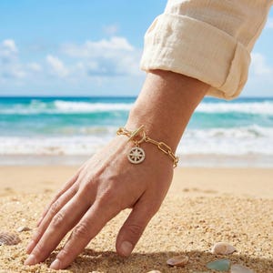 May include: A gold-toned chain link bracelet featuring a toggle clasp and a circular pendant with a starburst design encrusted with small clear stones. The bracelet is worn on a wrist, showcasing the metallic finish and the detailed charm.