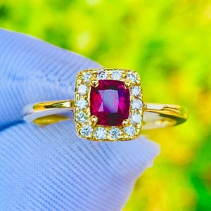 May include: A gold ring featuring a square-cut, deep red gemstone surrounded by a halo of small, clear gemstones. The ring's band is a polished gold color, and the setting is a square shape. The background is a blurred green and yellow.